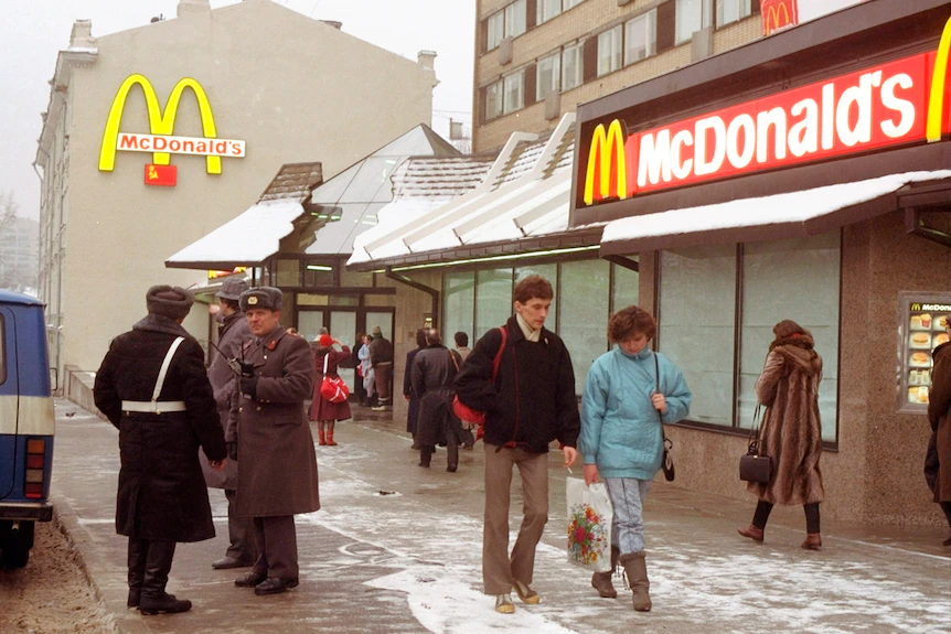 People walk past guards in Soviet-style military garb in front of a fast-food restaurant with red and yellow branding