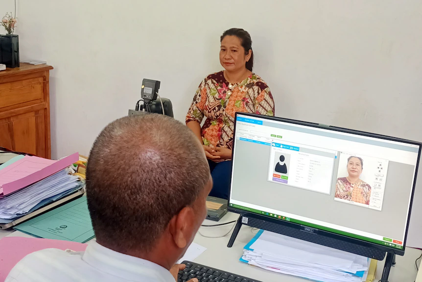 A woman sits for a driver licence photo while a man looks at the image on the screen 